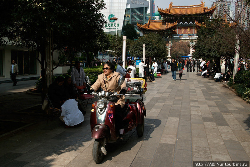 Популярное средство транспорта в Куньмине. На заднем плане, в белых халатах — уличные массажисты. Прямо посреди сквера стоят стулья, мануалы предлагают прохожим свои услуги. Куньмин, Китай